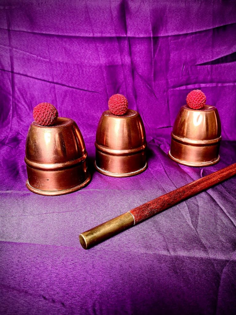 zusehen ist das Becherspiel aus Messing. Auf den drei Bechern liegen drei rote Bälle.